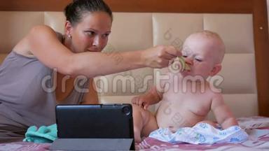 年轻的妈妈用一勺粥喂可爱的宝宝.. 宝宝坐在床上盯着平板电脑上的卡通片